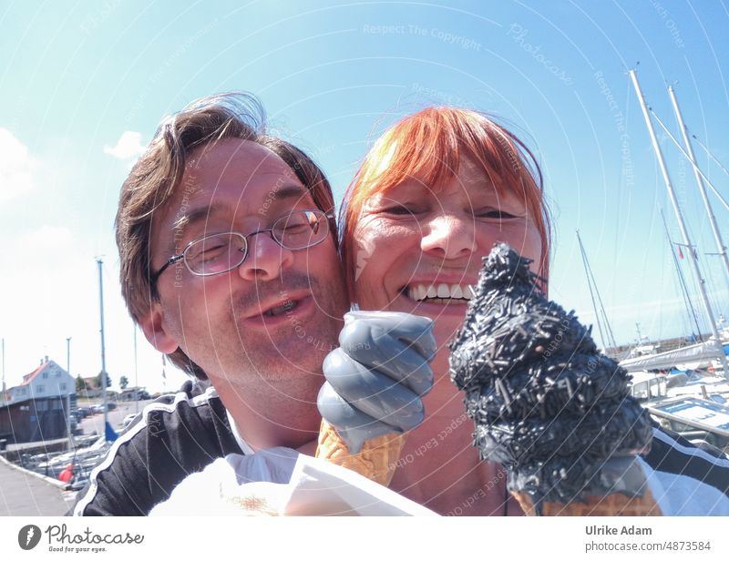 Lecker Lakritzeis |Glückliches Paar mit Eis in der Hand schaut freudig in die Kamera. Im Hintergrund sieht man einen Hafen bei schönem Wetter. Softeis glücklich