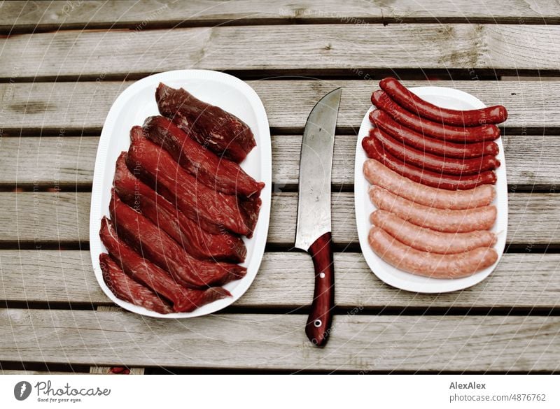 Ein Teller mit Rindfleischhüftsteaks und ein Teller mit Bratwürsten und Merguez- Würsten stehen nebeneinander auf einem Holztisch, in der Mitte liegt ein großes Fleischmesser mit Holzgriff.