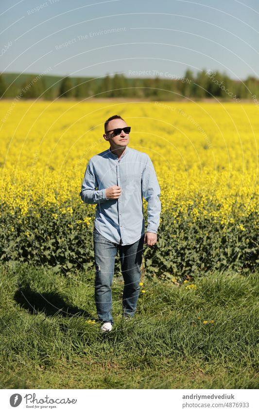 Ein junger Mann mit Sonnenbrille geht spazieren und amüsiert sich über ein sonniges gelbes Rapsfeld, das Konzept von Reisen und Freiheit. Sommerurlaubshintergrund
