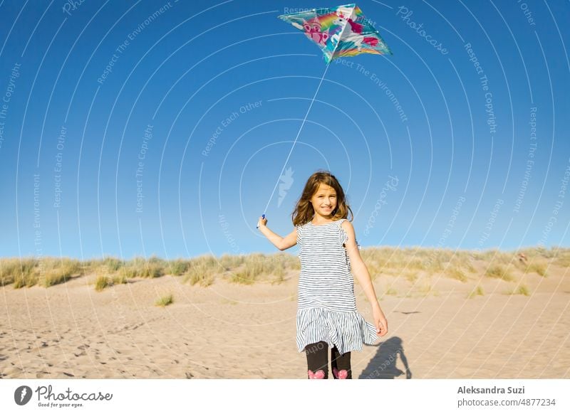 Nettes glückliches kleines Mädchen im Sommerkleid läuft mit fliegenden Drachen auf leeren Sandstrand. Schöner sonniger Tag, blauer Himmel. aktiv Aktivität