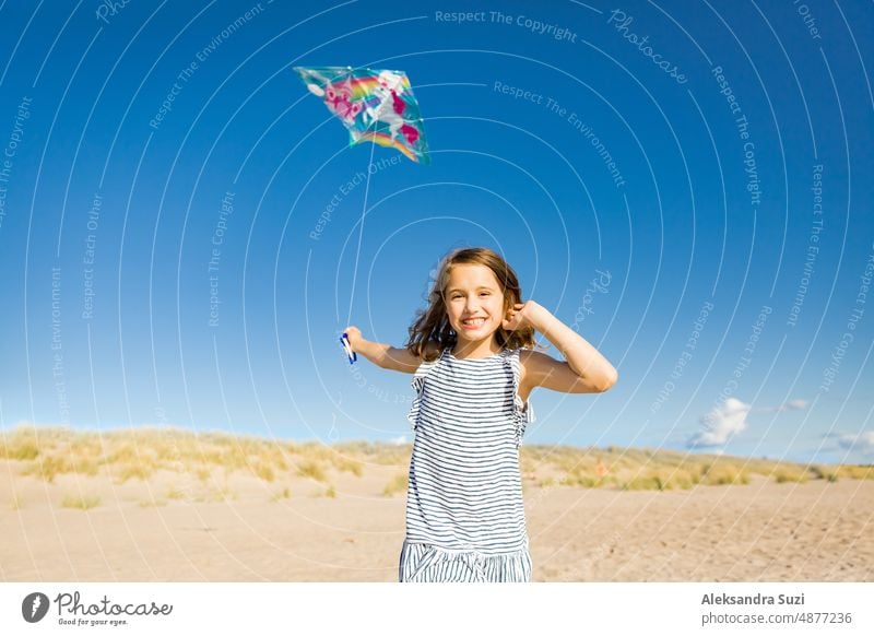 Nettes glückliches kleines Mädchen im Sommerkleid läuft mit fliegenden Drachen auf leeren Sandstrand. Schöner sonniger Tag, blauer Himmel. aktiv Aktivität