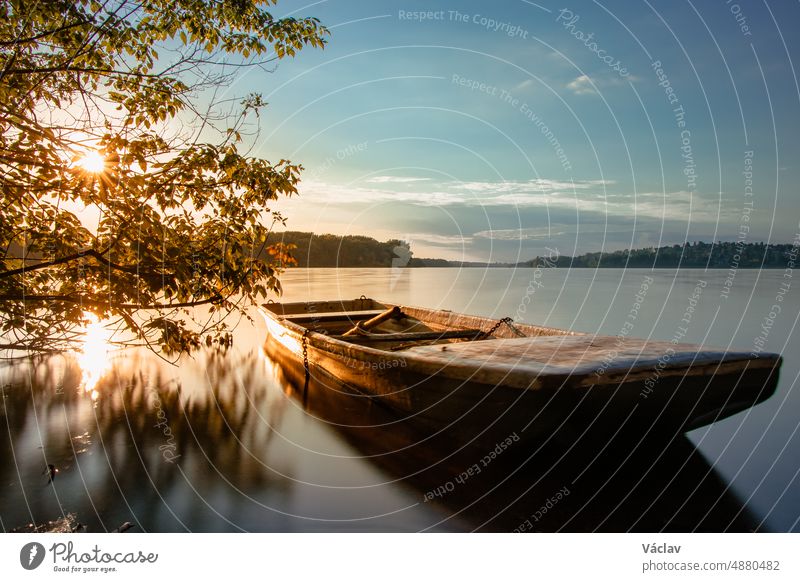Ein märchenhaftes Boot, das am Ufer der Talsperre Žermanice vertäut ist. Die untergehende Sonne, die durch die Bäume scheint, beleuchtet die atemberaubende Szenerie. Mährisch-Schlesische Region, Tschechische Republik