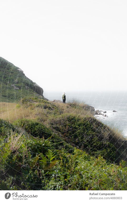 Mann mit Hut am Rande einer Klippe und Blick auf den Horizont vertikal Foto Fotografie grün Denken echte Menschen Landschaft weiß grau Strand Jahreszeiten