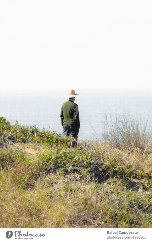 Mann mit Hut am Rande einer Klippe und Blick auf den Horizont Kontemplation Zeitgenosse Bildende Kunst weiß grau Strand Jahreszeiten Landschaft echte Menschen