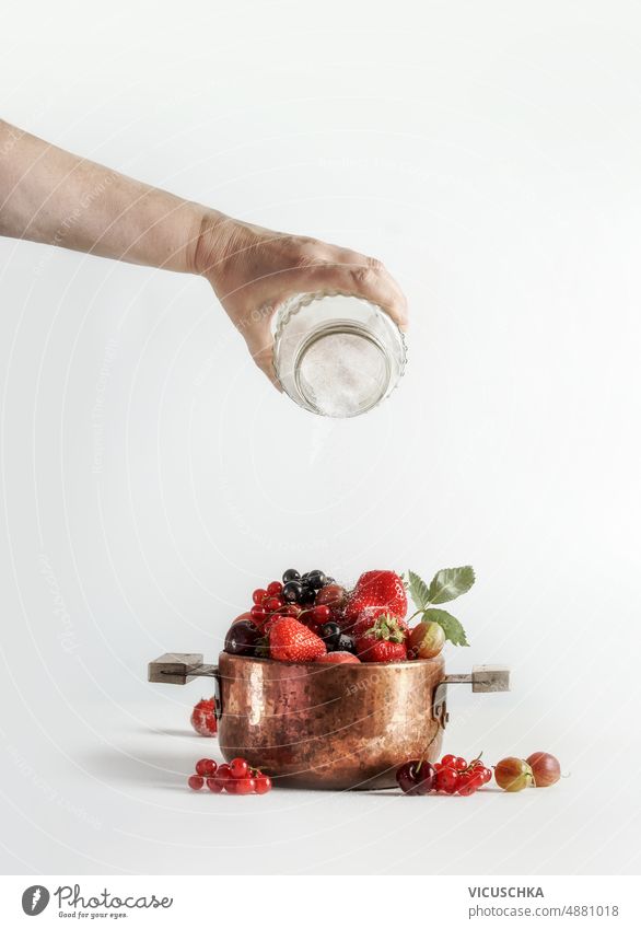 Marmelade Vorbereitung mit Frau Hand gießen Zucker auf Kupfer Kochtopf mit verschiedenen Sommer Beeren und Früchte auf weißem Hintergrund. Gießen kupfer