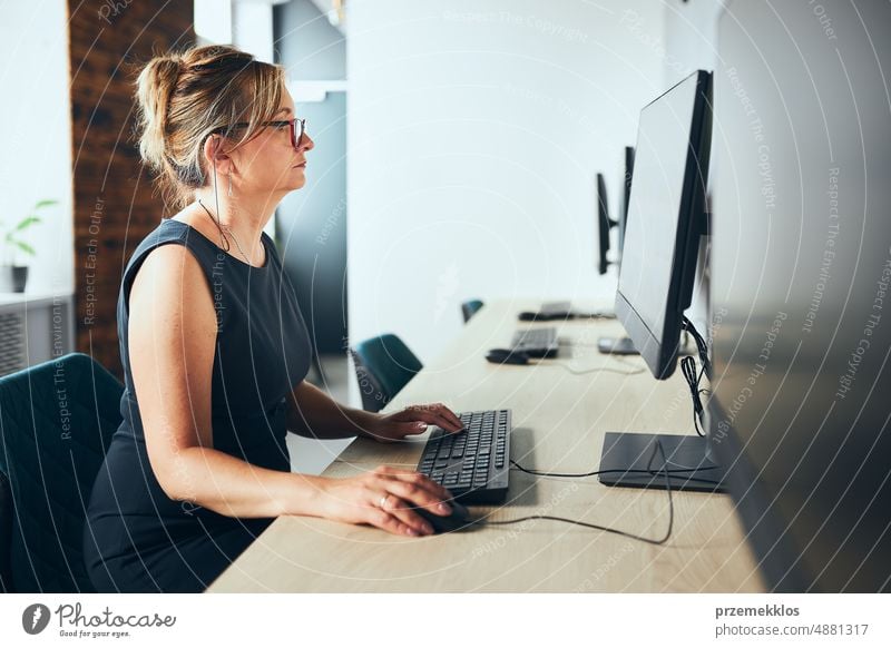 Besorgte Geschäftsfrau arbeitet am Computer im Büro. Beschäftigte Frau sitzt am Schreibtisch vor dem Computermonitor. Konzentriert auf die Arbeit. Offenes Büro. Kopierraum rechts