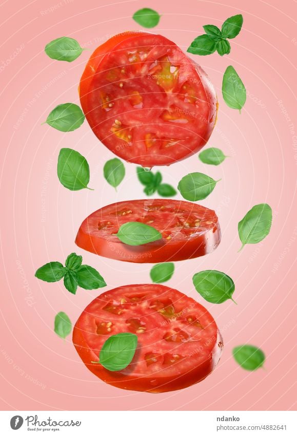 Runde rote Tomatenscheiben und Basilikumblätter schweben auf einem rosa Hintergrund. Gemüse Scheibe Lebensmittel Blatt Gewürz fallen grün Vegetarier Diät