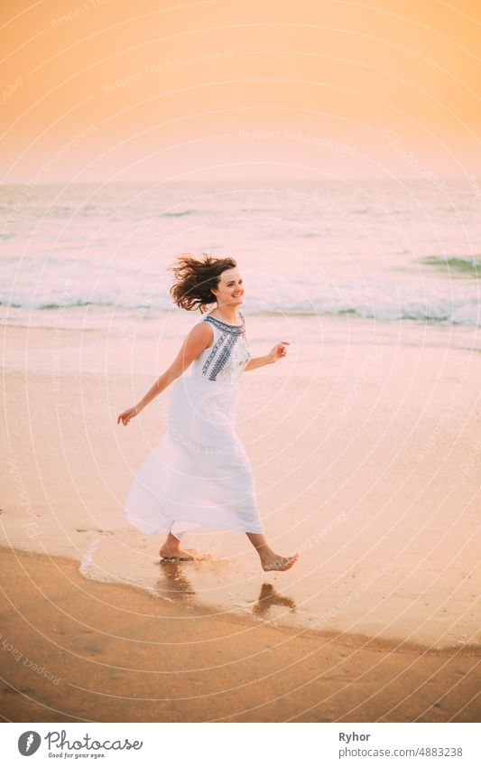 Goa, Indien. Junge kaukasische Frau mit flatternden Haaren im Wind im weißen Kleid, die am Meer entlang geht, das Leben genießt und im Sommersonnenlicht lächelt