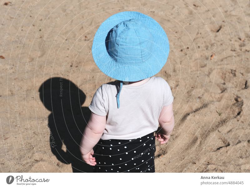 Ein kleines Kind mit blauem Hut bestaunt seinen Schatten Kindheit Kleinkind Außenaufnahme Spielplatz Sand sonnenschutz Farbfoto Aussenaufnahmen Schattenspiel