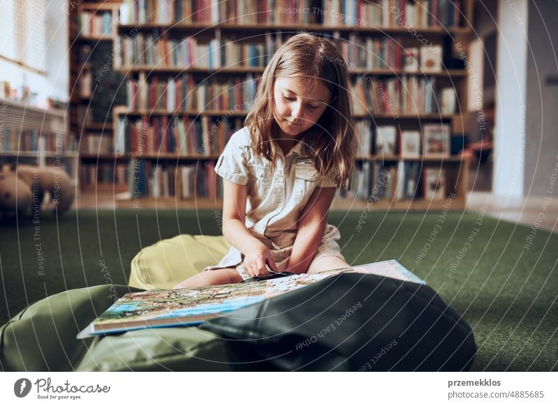 Schülerin beim Puzzeln und Lesen eines Buches in der Schulbibliothek. Grundschülerin beschäftigt sich mit einem Buch mit Puzzle. Vorteile des täglichen Lesens. Neugierde des Kindes