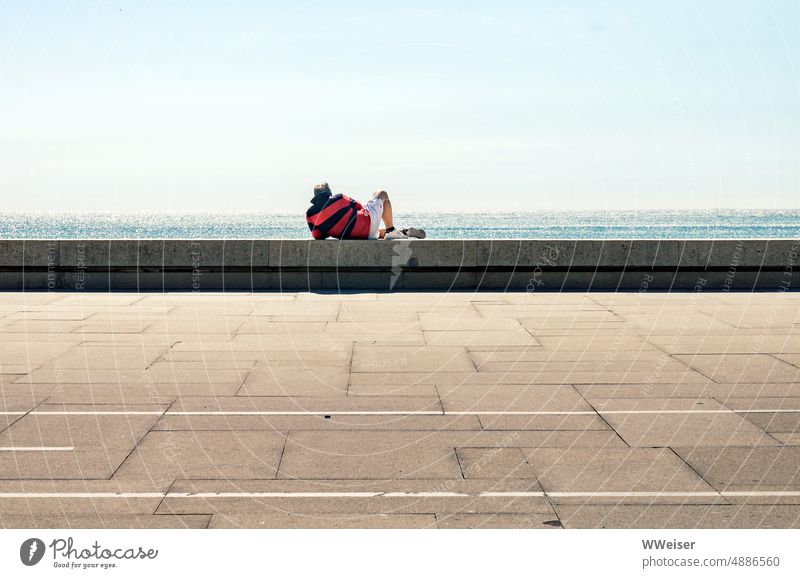 Ein Mann mittleren Alters liegt auf einer Mauer in der Sonne und schaut aufs Meer Küste Ozean Promenade Straße Pflaster liegen ausruhen entspannt Person Herr