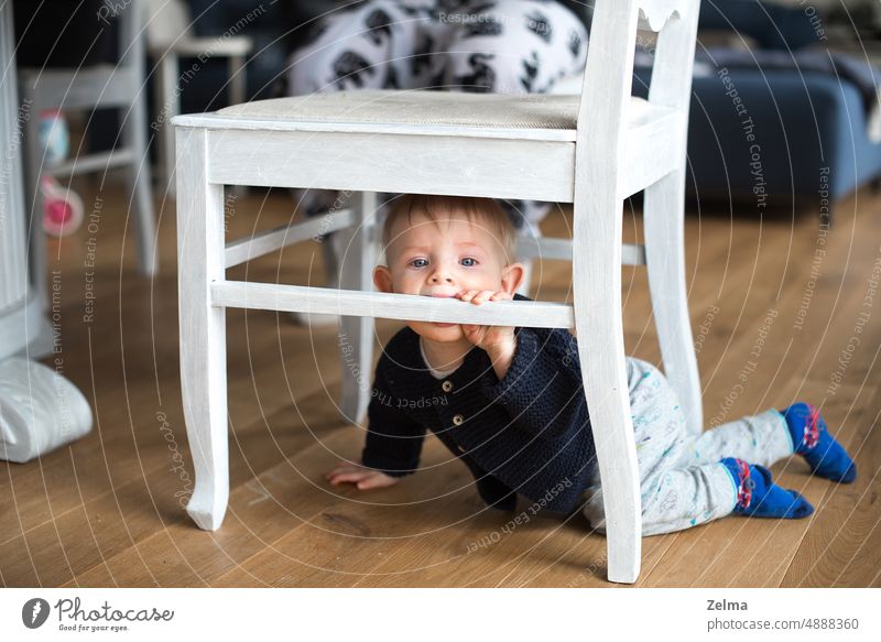 ein kleiner Junge spielt unter einem weißen Stuhl im Wohnzimmer der Familie niedlich Baby unten Spielen wenig Kind Kleinkind heimwärts im Innenbereich Glück