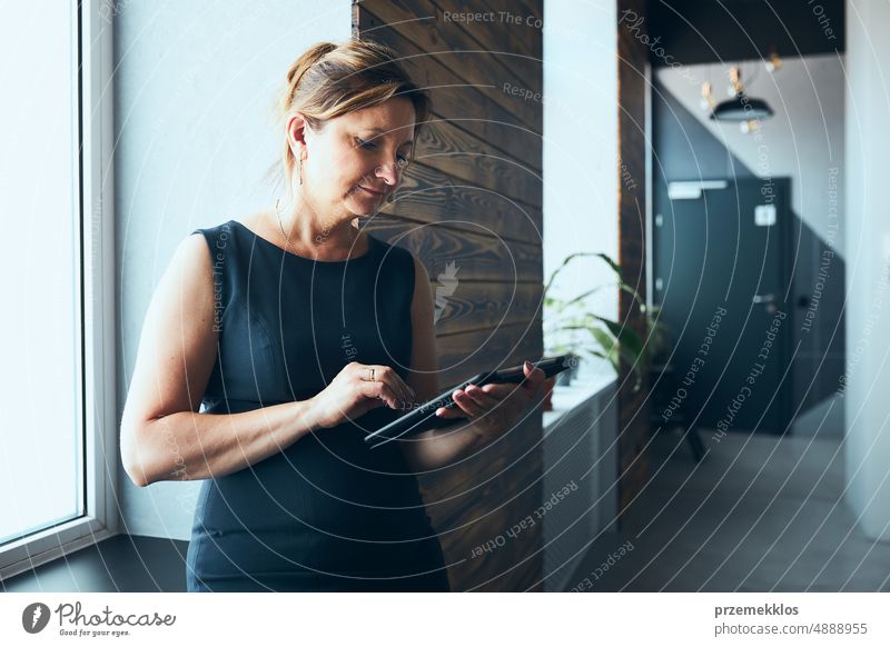 Geschäftsfrau arbeitet am Tablet im Büro. Reife Frau mit Touchpad-Computer stehend am Fenster in modernem Interieur. Manager konzentriert sich auf die Arbeit und hält ein digitales Gerät. Verwendung von Technologie