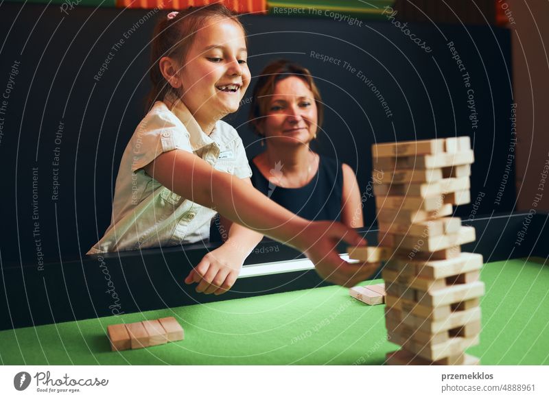 Aufgeregtes Mädchen spielt Jenga-Spiel mit ihrer Mutter im Spielzimmer. Mädchen entfernt einen Block vom Stapel und legt ihn auf die Spitze des Turms. Geschicklichkeitsspiel und Spaß. Familienzeit