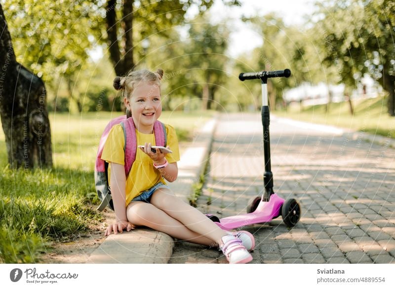 Ein reizendes kleines Mädchen mit einem Telefon in der Hand, mit einem Rucksack und einem Roller sitzt nach der Schule auf dem Bürgersteig. Zurück zur Schule, nach der Schule, Schulalter