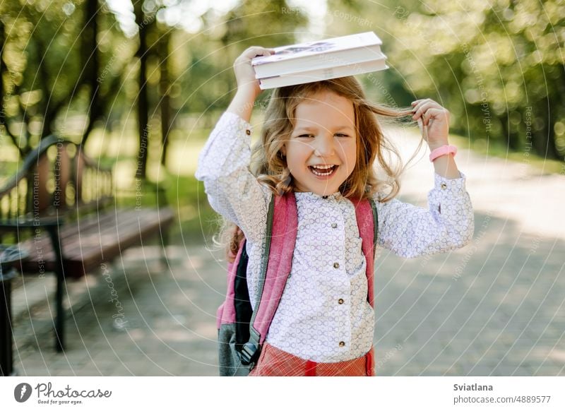 Ein bezauberndes kleines Mädchen mit einem Buch auf dem Kopf, am ersten Tag in der Schule oder im Kindergarten. Das Konzept von Bildung und Erziehung