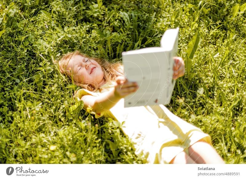 Ein bezauberndes Baby liegt mit einem Buch auf dem Rasen im Garten oder Park und blinzelt in die Sonne. Mädchen Schule Wissen Sommer ein Buch lesen Sommerzeit