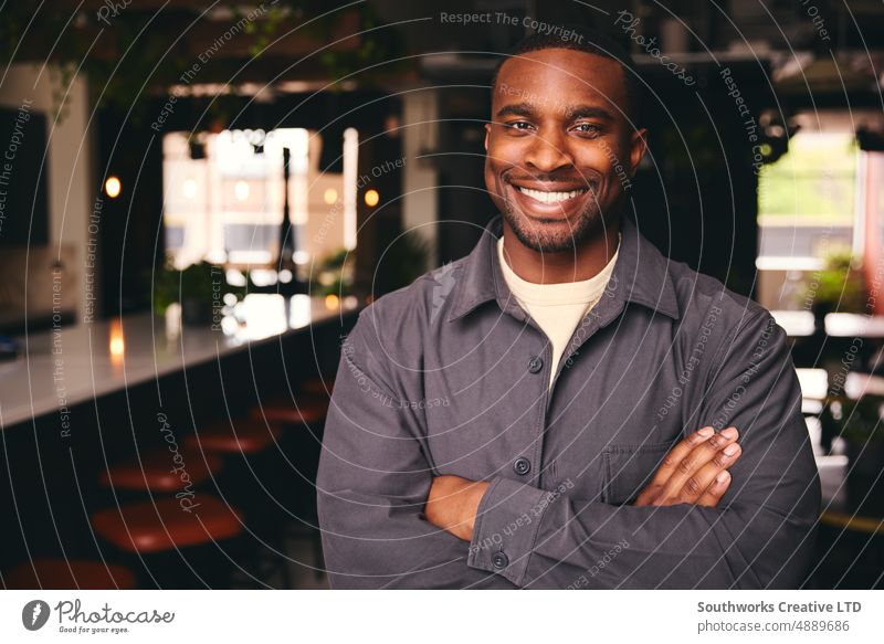 Lächelnder junger schwarzer Mann, der im Büro steht und mit gekreuzten Armen in die Kamera schaut selbstbewusst männlich professionell Kaffeehaus Afroamerikaner