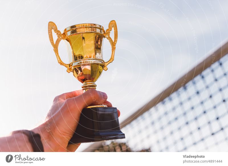 Goldener Pokal in der Hand des Sportwettkampfsiegers mit Spielnetz im Hintergrund Tennis Gewinner Tasse golden Preis Raster Volleyball Badminton Griffe Kelch