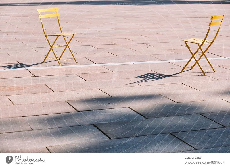 zwei leere gelbe Klappstühle stehen sich auf einem öffentlichen Platz gegenüber und werfen lange Schatten Stühle Sonne sonnig einladend Stadt urban Klappstuhl