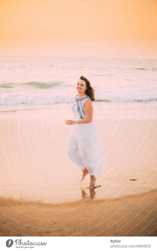 Goa, Indien. Junge kaukasische Frau in weißem Kleid zu Fuß entlang Meeresufer, Blick auf Kamera und lächelnd im Sommer Sonnenlicht Arabisches Meer