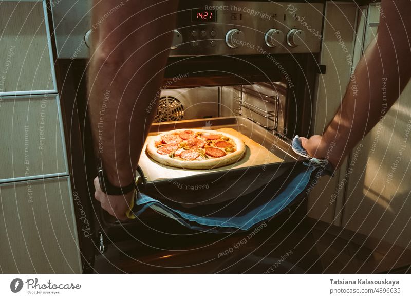 Ein Mann backt zu Hause eine Pizza im Ofen. Männerhände ziehen ein Pizzabackblech aus dem Ofen. männlich Hände Koch machen backen Peperoni Jalapeno Chilischote