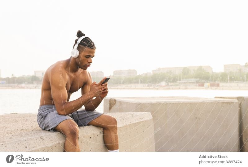 Schwarzer Sportler surft auf seinem Smartphone, während er an der Strandpromenade Musik hört MEER meloman Gesang Melodie Browsen online Internet Funktelefon