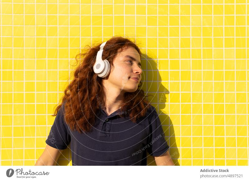 Frau mit Kopfhörer an der Wand stehend Headset Wegsehen Rotschopf gelb Sonnenlicht Stehen Fliesen u. Kacheln T-Shirt lockig Behaarung Musik Lifestyle