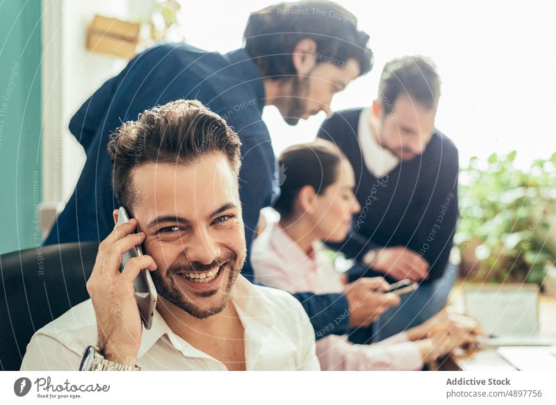 fröhlicher Mann, der in der Nähe von Kollegen mit seinem Smartphone spricht Arbeiter Telefonanruf Gespräch diskutieren Business Büro Mitarbeiterin