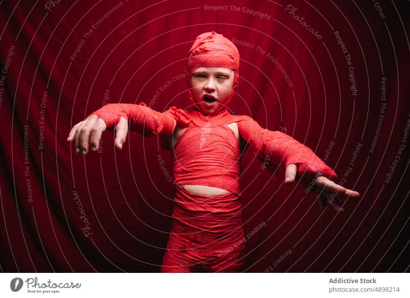 Niedliches kleines Kind im Mumienkostüm, das im roten Studio schreit und gestikuliert Halloween Tracht Junge beängstigend Porträt spukhaft sich[Akk] melden