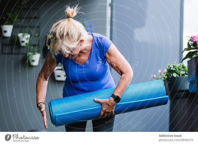 Aktive ältere Frau mit gerollter Yogamatte im Fitnessstudio Senior Unterlage aktiv Gesundheit Lifestyle Beteiligung blau smartwatch vorbereitend Training