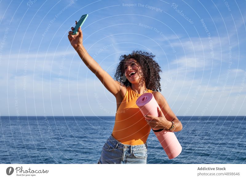 Fröhliche hispanische Frau mit Matte macht Selfie am Meer Yoga Unterlage Ufer MEER Gesunder Lebensstil Training Hobby Selbstportrait soziale Netzwerke