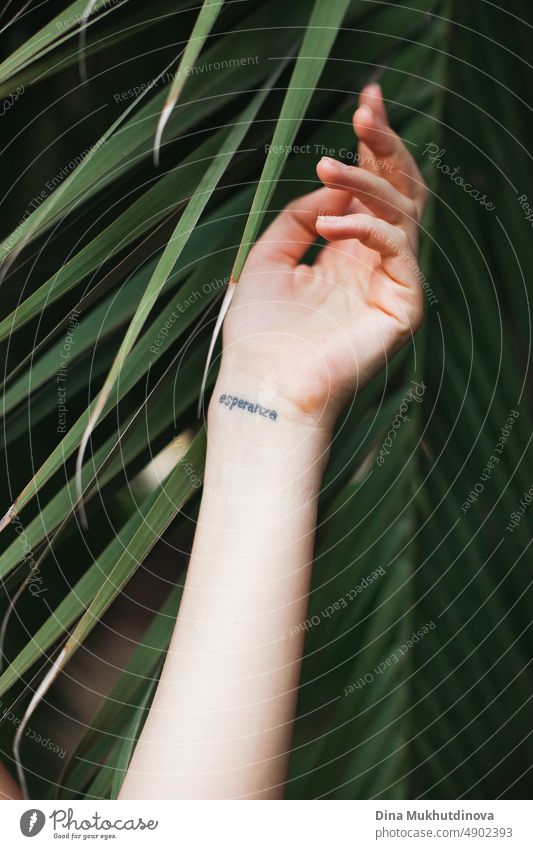 Hand entspannt mit grünem Palmblatt als Hintergrund mit Wort "Hoffnung" in Spanisch. Tattoo-Kunst auf einem Handgelenk der Hand einer Frau im Sommer. Berührende tropische Natur. Konzept des Zusammenlebens von Natur und Mensch. Umweltfreundliches Konzept. Tropische Schönheit.