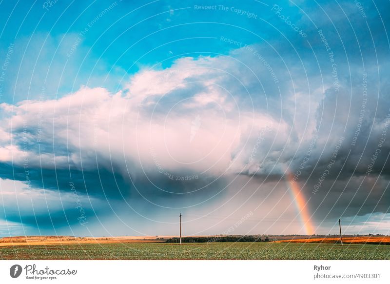 Dramatischer Himmel während des Regens mit Regenbogen am Horizont über ländlichem Landschaftsfeld. Landwirtschaft und Wettervorhersage Konzept. Ländliche Wiese im Herbst regnerischen Tag