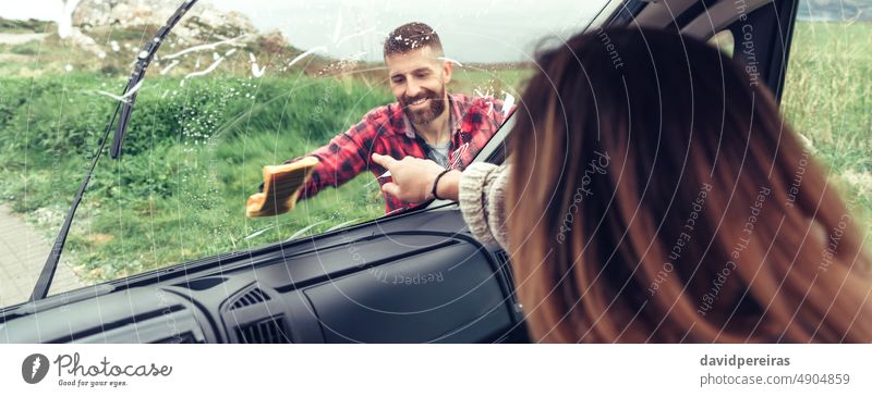 Mann reinigt die Windschutzscheibe eines Lieferwagens, während die Frau auf einen Fleck zeigt Lächeln Reinigen Spülung Glas Kleintransporter Transparente Netz