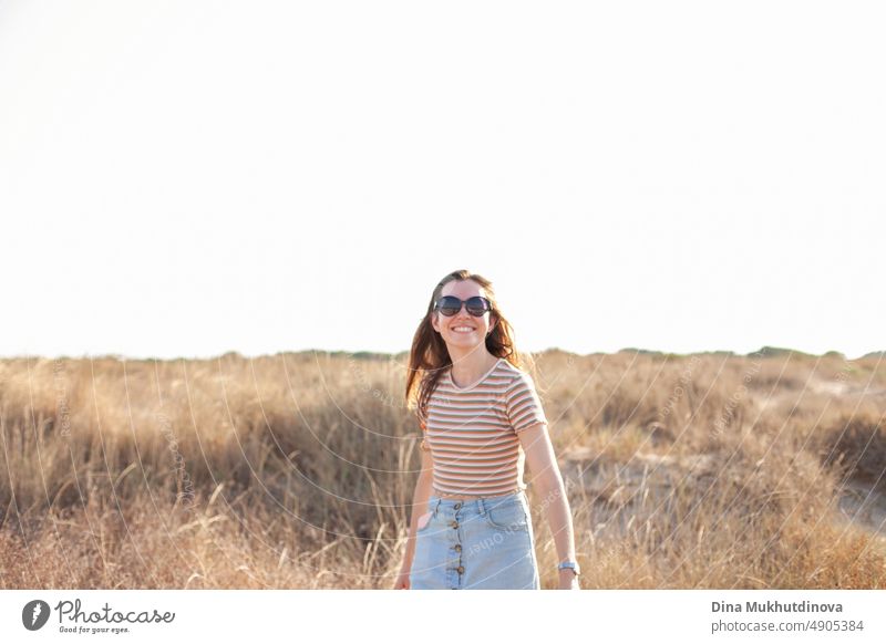 Glückliche junge Frau mit Sonnenbrille, die in der Nähe des Strandes in den Dünen mit hohem trockenem Gras spazieren geht. Foto in neutralen Beigetönen.
