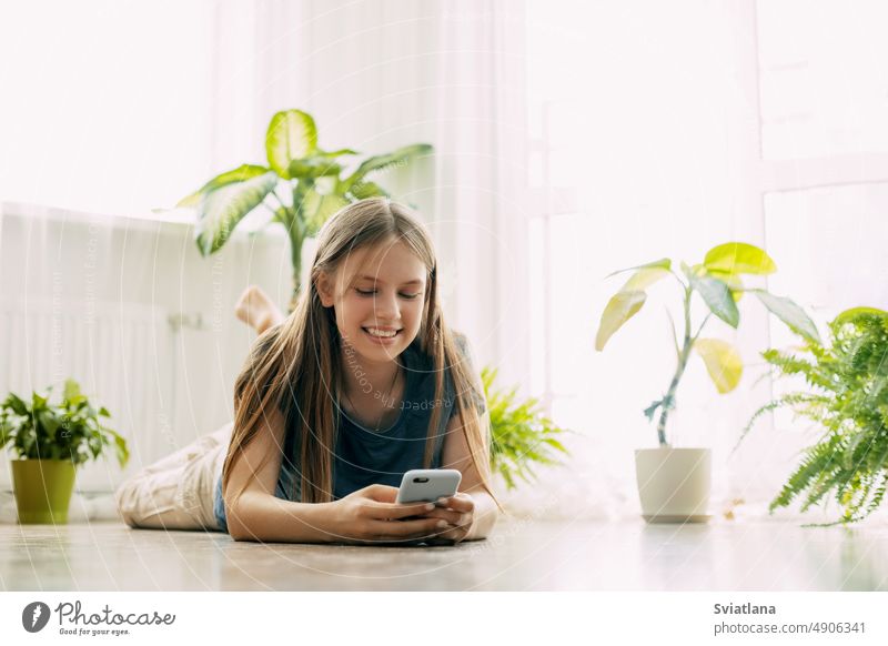 Ein glücklicher Teenager schreibt eine SMS auf seinem Handy, das auf dem Boden im Wohnzimmer liegt. Moderne Technologien, Online-Kommunikation Schüler Mädchen