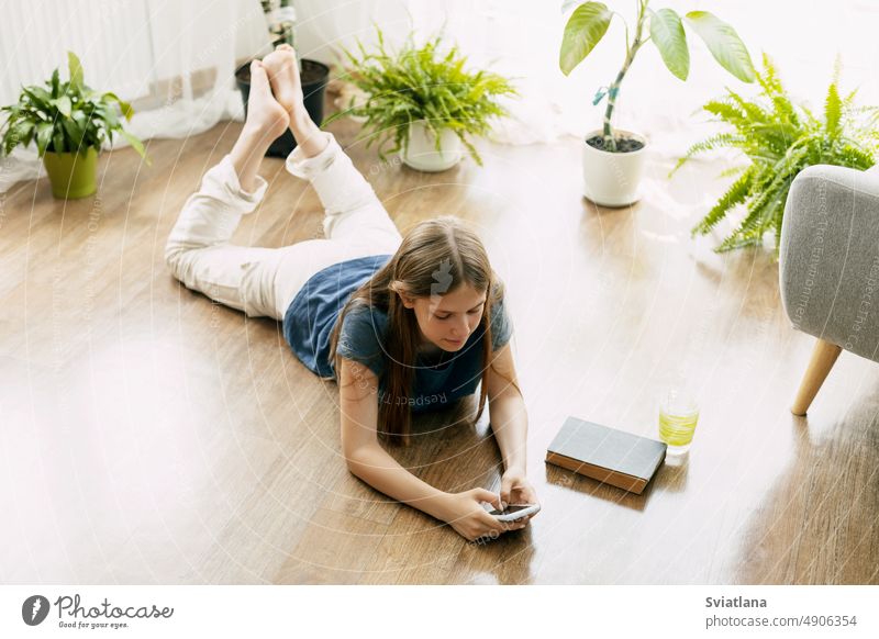 Ein glücklicher Teenager schreibt eine SMS auf seinem Handy, das auf dem Boden im Wohnzimmer liegt. Moderne Technologien, Online-Kommunikation Schüler Mädchen