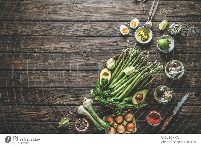 Lebensmittel Hintergrund mit grünem Spargel und verschiedenen gesunden Zutaten: Avocado, Ei, Frühlingszwiebel, Limette, Gewürze und Kräuter auf dunklen rustikalen Holztisch Küche