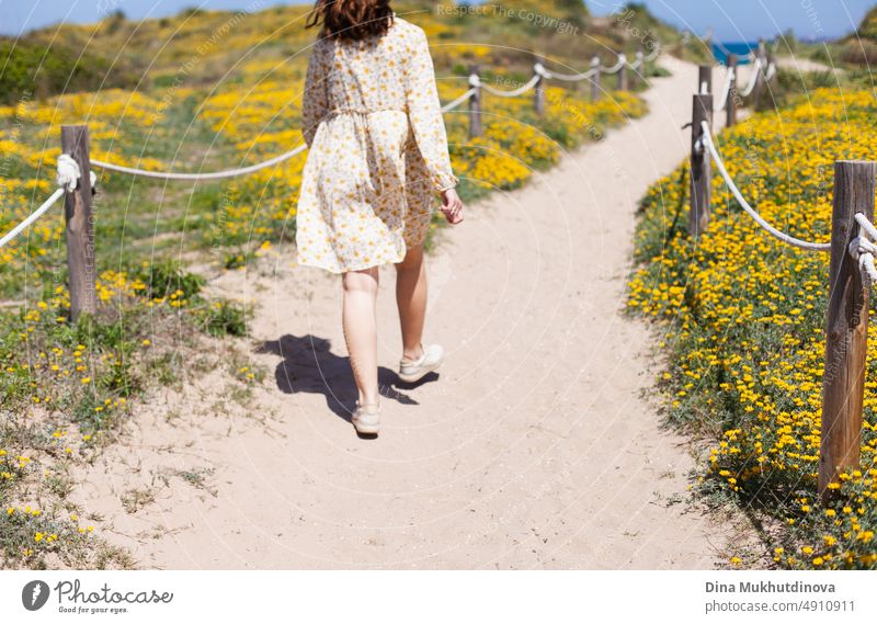 Junge Frau im Sonnenkleid von hinten am Strand zwischen blühenden Dünen mit gelben Blumen. Foto in neutralen Beigetönen. Reisender reisen schön Wahrzeichen