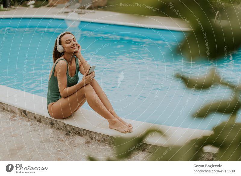 Junge Frau sitzt am Swimmingpool und hört Musik vom Mobiltelefon mit Kopfhörern im Hinterhof des Hauses Bikini Kaukasier Tag Telefon genießend Außenseite frisch