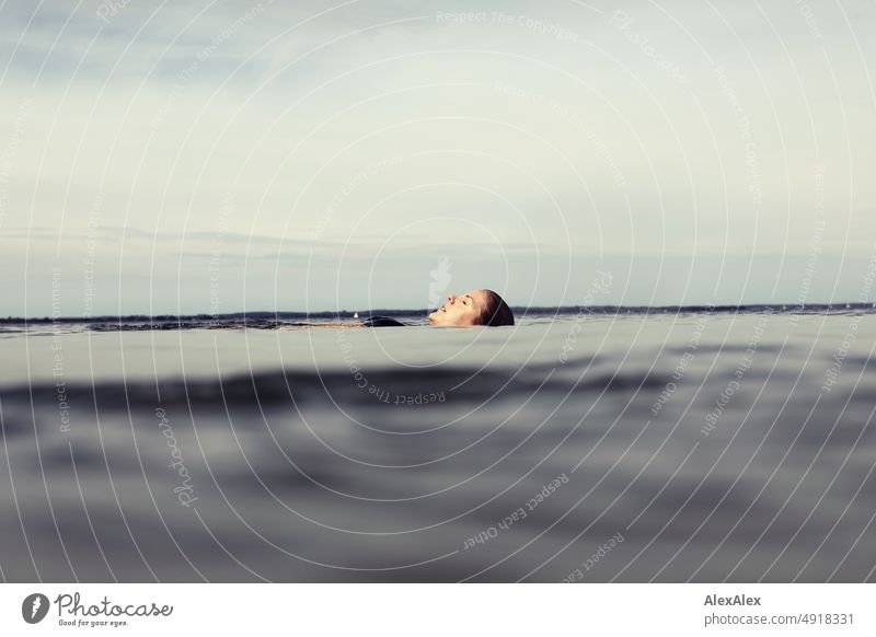 Junge, schöne Frau liegt im flachen Wasser der Ostsee und treibt liegend mit geschlossenen Augen auf der ruhigen See Lifestyle Gegenlicht gesund sportlich