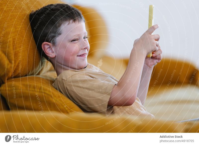 Glücklicher Junge sitzt auf einem Sofa und surft auf seinem Smartphone Kind heimwärts Telefon Browsen Lächeln Freude Technik & Technologie jung konzentriert