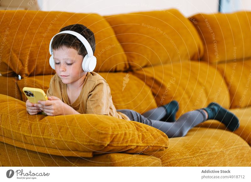 Auf einem Sofa liegender Junge, der ein Mobiltelefon benutzt Kind Lügen heimwärts Handy Technik & Technologie Smartphone Kopfhörer jung zuhören ernst