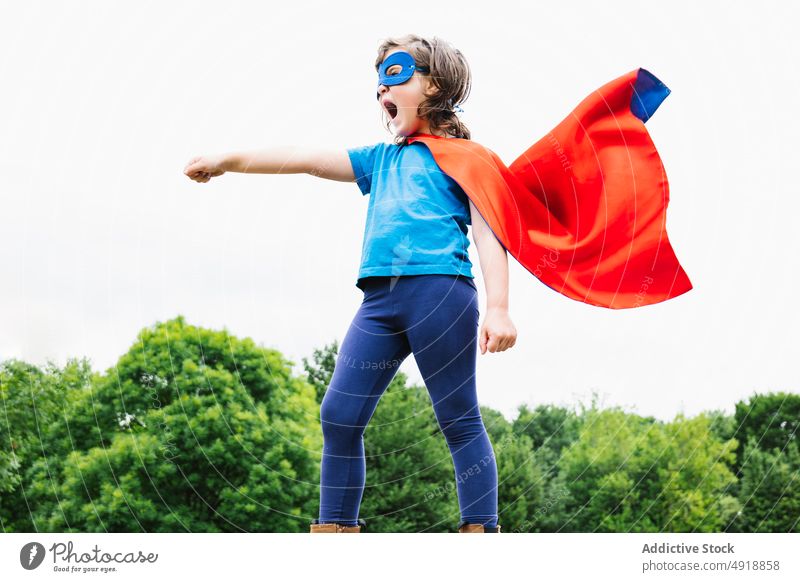 Aufgeregtes Mädchen im Superheldenkostüm im Park schreien spielen Baum Tracht Arm angehoben Held Kraft wolkig Frau Kap Mundschutz aufgeregt Sommer Kind tagsüber