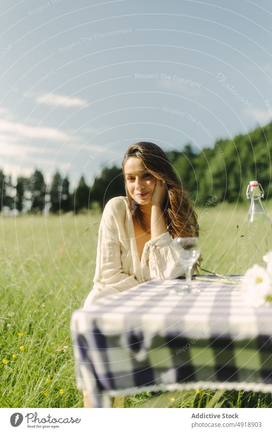 Positive Frau sitzt am Tisch in einem Feld Picknick Landschaft Sommer Erholung Natur Gras Zeitvertreib ruhen feminin Lächeln Getränk Kleid Optimist Inhalt