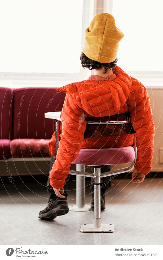 Unerkennbare Frau in Oberbekleidung auf einem Stuhl ruhend Raum Fenster Sofa sonnenbeschienen fettarm Stil Island Mantel Beanie Hut Sonnenlicht lässig Kälte