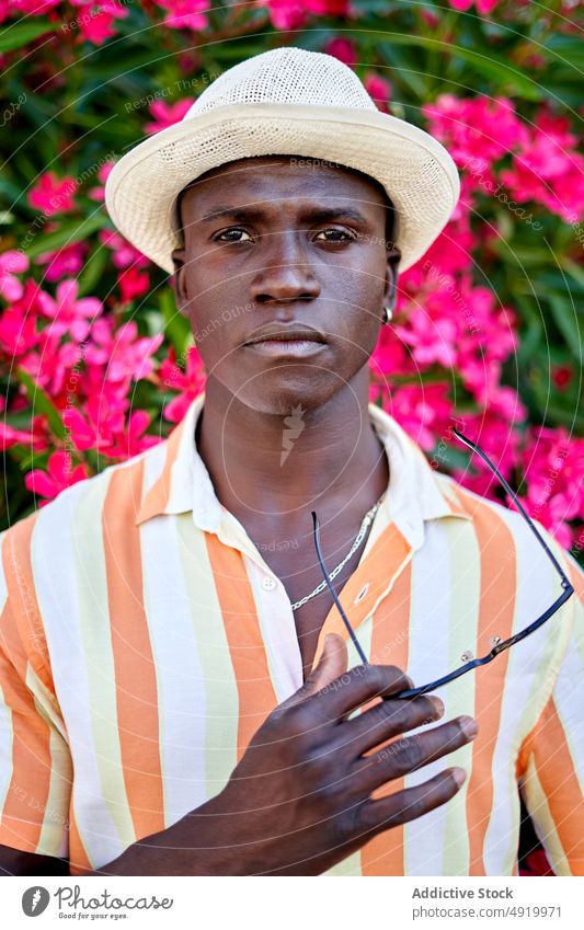 Ernster schwarzer Mann in der Nähe des blühenden Busches Straße Buchse Blume Pflanze Vorschein Großstadt geblümt Kleidung Stil Afroamerikaner ethnisch Sommer