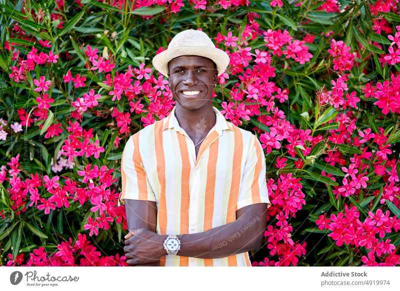 Glücklicher schwarzer Mann in der Nähe eines blühenden Busches Straße Buchse Blume Pflanze Vorschein Großstadt geblümt Kleidung Stil Afroamerikaner ethnisch
