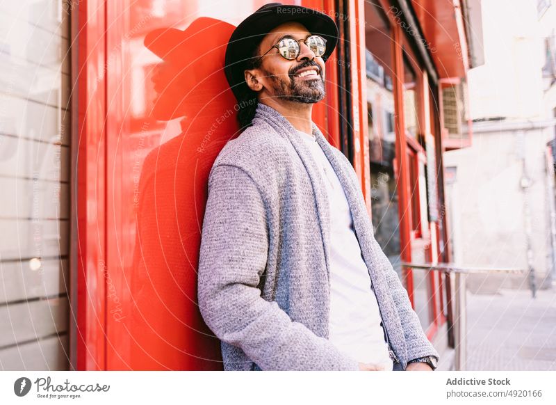 Fröhlicher hispanischer Mann, der sich an die Wand lehnt fettarm Straße Lächeln Gebäude Glück Streetstyle urban ruhen männlich Erwachsener ethnisch Stil trendy